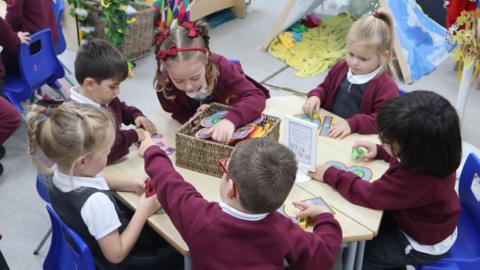 Six young children, three boys and three girls wearing burgundy school jumpers and grey school dresses, sit around a hexagonal shaped table on blue plastic chairs doing crafts. 
