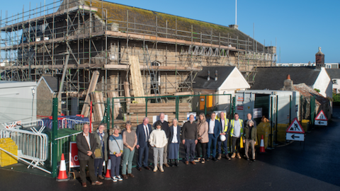 A wide shot of the town hall. It is an old stone building, mainly obscured by scaffolding. Various construction gear is around the building, including signs, fences, and cones. 15 people are stood in a line in front of the building. 