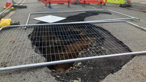 A hole in the ground covered by grey fencing, which is enclosed by cones and other barriers