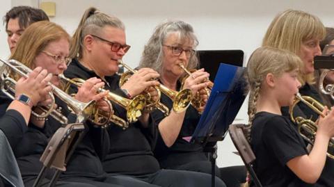 Five woman and a girl are dressed in black playing brass instruments.