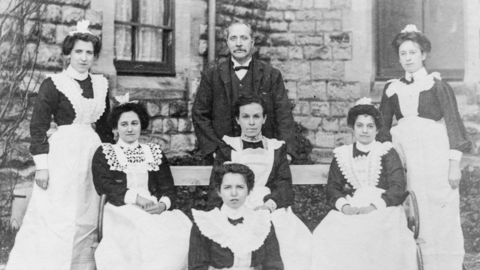 Three rows of women wearing Victorian hospital uniforms - dark dresses and white aprons - and a man are pictured sitting or standing in front of a stone building.