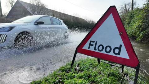 A car driving through floodwater on a country road, beside a flood warning sign