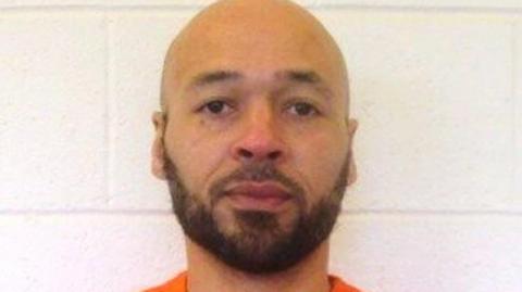 A mug shot of Tremane Wood wearing an orange prison shirt as he stands against a white wall