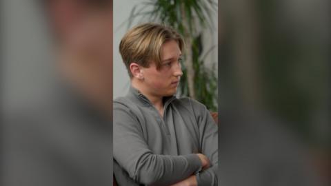 Max Murray sits with his arms crossed. He has blond hair styled as curtains and wears a grey top 