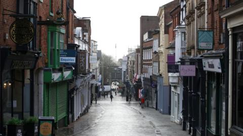 A view of a shopping street looking downhill. There is a row of shops on either side of the cobbled street. The shops are closed with shutters down on a few of them.