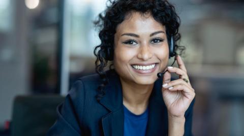 A woman wearing a dark blue blazer and blue t-shirt and a headset smiles towards the camera, seemingly during a call