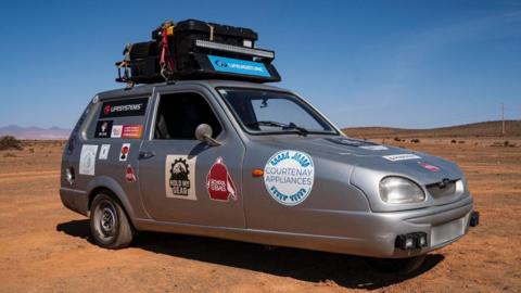The Reliant Robin in the middle of the desert. The three-wheel vehicle is silver and has stickers on the side of it and on the bonnet.
