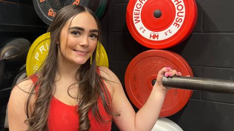 Annie, stood next to a weight rack wearing a red gym set. She has lots of different weights behind her as she smiles at the camera. 
