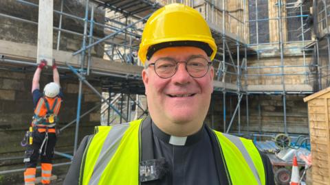 A head and shoulders photo of a man in a yellow hard hat, with a black top and clerical collar on and high vis vest. He is wearing black rimmed glasses and smiling. Behind him you can see scaffolding with a workman in orange high vis lifting up a wooden panel.