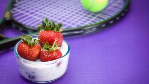 A pot of strawberries and cream next to a tennis racket and a tennis ball.