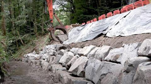 Rows of large rocks arranged on a bank with the arm of a mechanical digger seen moving one of them. Woodland can be seen in the background.