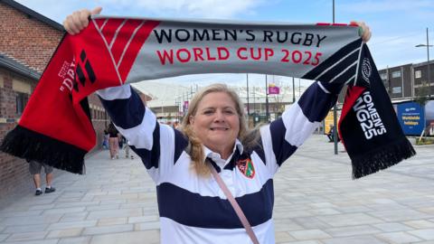 A woman wearing a striped white and blue rugby shirt holding her arms aloft and smiling. She is holding a banner proclaiming "women's rugby world cup 2025". Behind her the roof of a stadium can be seen.