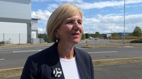 A woman with short blonde hair styled in a bob is standing on a pavement next to a road and a roundabout. She is wearing a black blazer jacket and a white top, with a black and white badge pinned to her lapel.