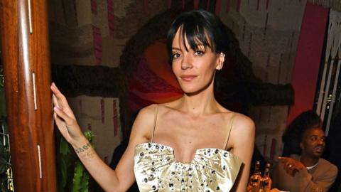 Lily Allen, looking to camera, wearing a gold strap dress, with her hand on a wooden pole.
