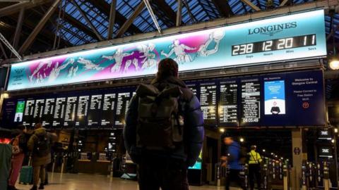 A man with a backpack standing in a railway station, looking up a the large departure boards and the countdown clock of the Commonwealth Games - a blue and purple designed illustration of athletes playing sport, with a clock ticking down to the Games starting.