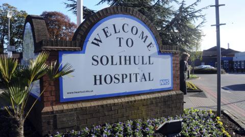 A brick structure has a white a blue sign mounted to it that reads "Welcome to Solihull Hospital" with small plants at the base. 