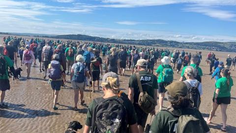 A large crowd of several hundred people is walking together across a wide sandy beach at low tide on a bright, sunny day. The photograph is taken from behind the group, looking toward the sea and a distant shoreline.