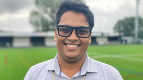 Rohan stands on the pitch at Brigg Town and smiles for the camera.  He wears a thick pair of black glasses and a light blue shirt.