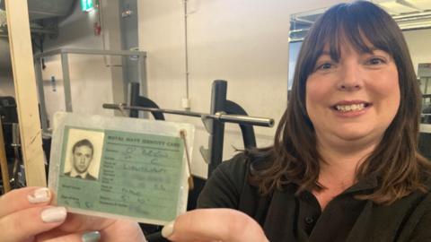 Morwenna Philpott is smiling as she holds the identity card up to the camera as she strands in the gym. She has long dark hair and is wearing a black top. The card has a head and shoulders shot in black and white photo of a man wearing a white shirt, dark jumper and tie. It is labelled ROYAL NAVY IDENTITY CARD and in biro is written: O'Brien, PC, Lieutenant, Date of Issue 16.9.81, Height 5'10". The date of birth is partly obscured and the service number is totally obscured. 