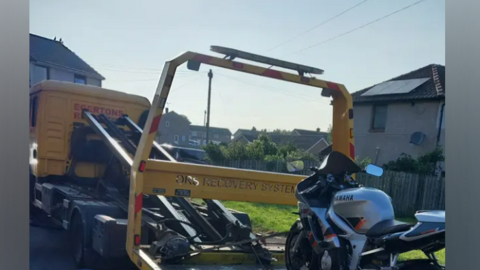 A Yamaha silver-and-black motorbike being loaded onto a pick-up truck on a housing estate on a sunny day.