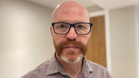 A head-and-shoulders picture of John Taylor, who is wearing glasses and is looking at the camera. He has a ginger beard flecked with grey and is wearing a brown checked shirt. The background, a room with plain cream walls and a wooden door, is out of focus.