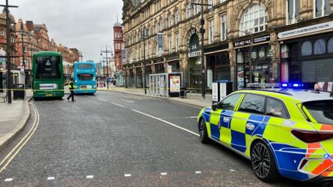 A road through a city centre with shops on either side. Part of the road has been cordoned off with yellow police tape. In the cordon there are two stationary double decker buses. Parked outside of the cordon is a police car and a police officer is stood behind the cordon. 