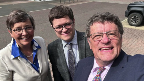 A man with dark grey curly hair and in a navy blue suit, purple shirt and floral tie takes a selfie with a woman with short dark hair wearing a beige jacket and blue blouse alongside a young man with dark hair and wearing a grey suit. The three are stood in a car park on a sunny day.
