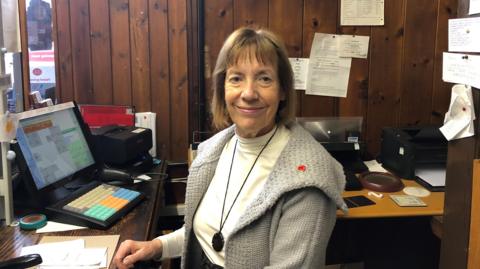 A woman in a grey coat with a Remembrance Day badge smiling at the camera. She si behind a counter with a computer and wooden walls.