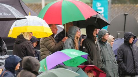 A crowd of people at the Royal Cornwall Show in Wadebridge hold up multi-coloured umbrellas during a shower. One of the umbrellas is from the Wimbledon tennis championships. Several people are wearing coats with hoods up.