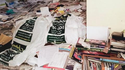 A bedroom full of hoarded items and old papers. The bed is not visible and each surface is covered with piles of items