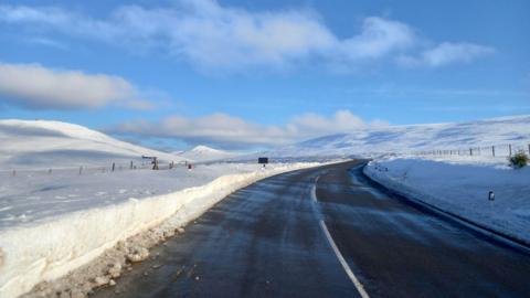 The Mountain Road, which which has been cleared but has snow either side. There are snow covered hills in the distance.