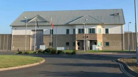 The exterior of Jurby Prison, a large brown building, with flag poles and CCTV cameras, there is a road in front and grass on either side.