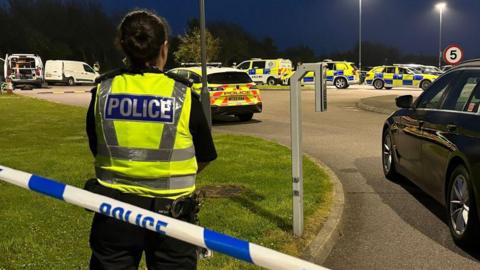 Back of female police officer in yellow hi-vis jacket with word 'police' on it in silver and blue, behind blue and white police tape, with multiple police vehicles with blue and yellow checks, at the scene of a fatal accident in a car park.