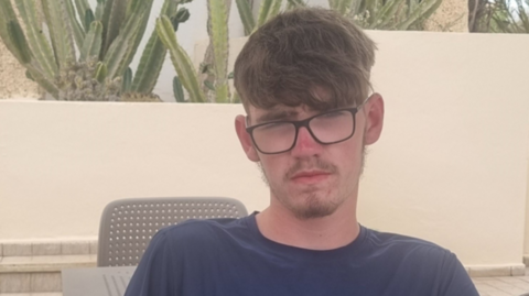 A young man with thick dark hair, facial hair and black framed glasses. He is wearing a blue t-shirt.