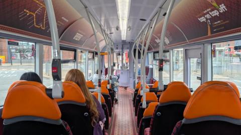 View is from the back of a bus, with the gangway down the middle. The seats either side have orange headrests and there are handle rails down either side. Windows fill each side of the bus and there are two women sat together on one set of seats, one brunette and one blonde and they have their backs to the camera.