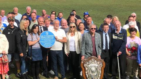 A group of about 40 people standing on a football pitch. In front a pair hold a blue plaque while a man holds a silver shield on a wooden background. 