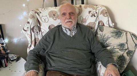 Michael Heelas sat in his living room at Turville Court, Great Bookham. Heelas is wearing a green jumper and brown trousers. He has white hair and a short white beard.