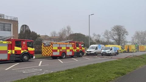 Fire crews and ambulances parked on the road outside an industrial area. It is cloudy or foggy. There is the frame of a green-coloured petrol station in the background.