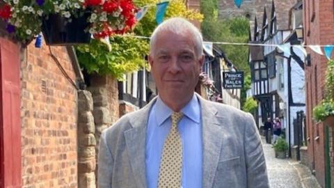 Ewen Sinclair wearing a grey jacket, blue shirt and a yellow and blue dotted tie. He has grey hair and is smiling at the camera. He is in a narrow alleyway and there are old buildings behind him.