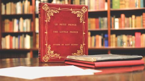 A red-covered book stands on a table with the words written on it: Le Petit Prince, The Little Prince, Antoine De Saint Exupery. Pieces of paper and a thin, old book is on the table in front of the red-covered book.