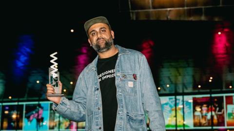 Aly Jamal wearing a black t-shirt and blue denim jacket, and a hat. He is looking at the camera and holding the award which is white and silver. 