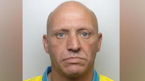 A bald man in his 40s, wearing a yellow top with a blue collar which looks like a football shirt. He is staring into the camera.