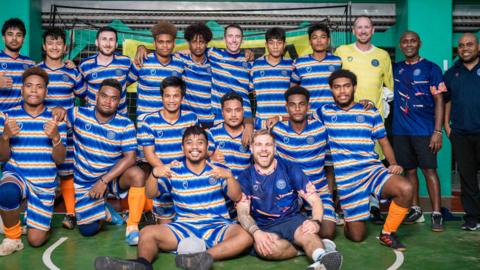 This image captures a team photo taken on an indoor sports court. Nineteen individuals are posing together, with some standing and others kneeling or sitting in the front row.
They are all wearing matching uniforms: blue, orange, and white striped shirts with orange socks.
In the background, there's a goal net, suggesting this could be a futsal or indoor football team.
The walls are painted in bright colors—green, orange, and blue.