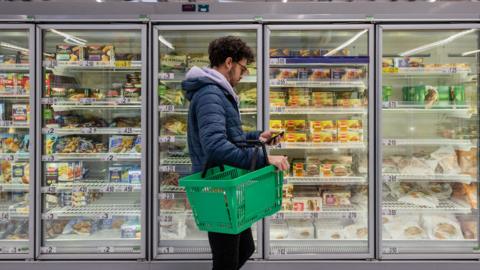 A man shopping in a supermarket, standing in front of the big fridges with a shopping basket.