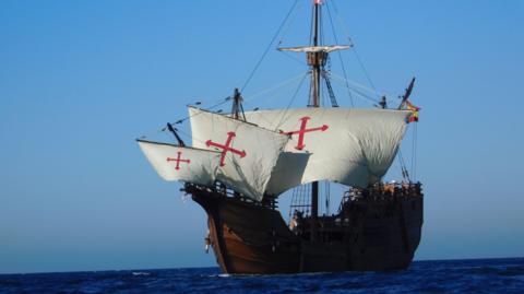 A replica of Christopher Columbus's flagship, The Nao Santa María. It is a tall wooden ship with four masts, five sails, and five decks. The sails are white with a red cross. It is sailing.