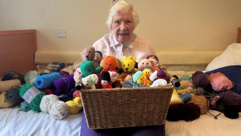 The picture shows Peggy sitting her room at Brunelcare. She is holding a basket containing her knitted creations.  She is surrounded by balls of wool.