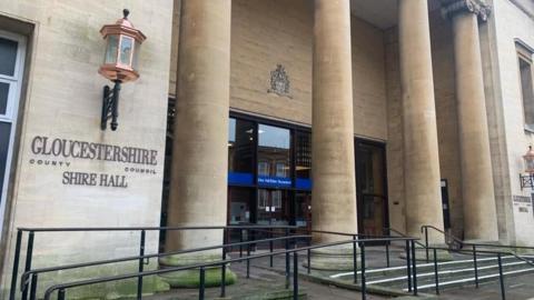 The exterior entrance to Shire Hall in Gloucestershire. It shows a sand coloured building with four large pillars outside, and a large sconce lantern. 