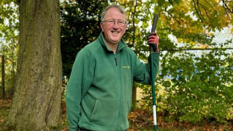 John Eggleston who is smiling at the camera. He has grey hair and is wearing a green fleece and gardening gloves. He's standing in a garden.