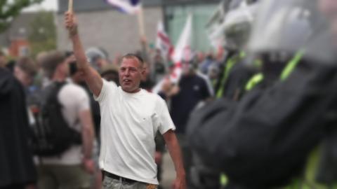 An image of Stephen Roughley in a white t-shirt, holding an item aloft in his right hand at a riot. 