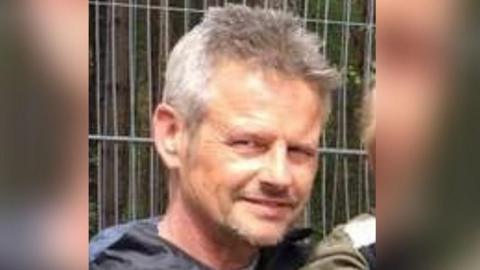 Chris Davies smiles at the camera. He has short grey hair and is wearing a dark coloured top. He is standing in front of some metal fencing.